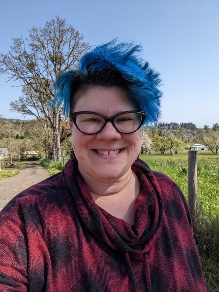 smiling woman with short blue hair wearing dark-rimmed glasses on a sunny day