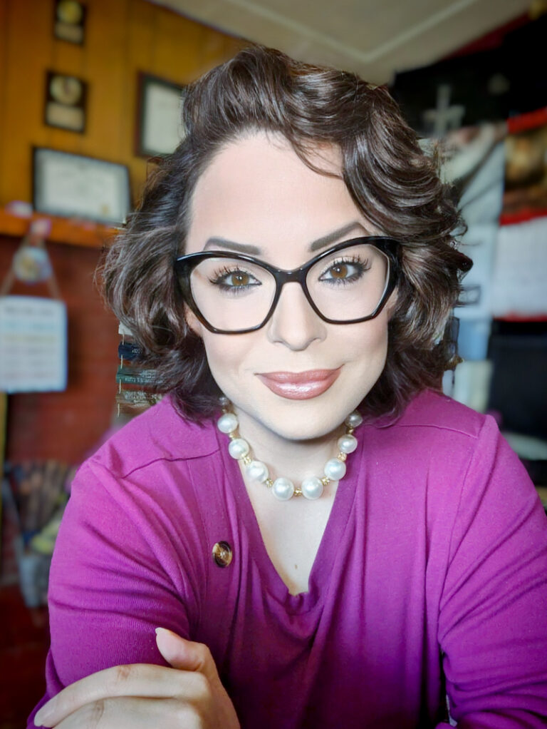 Female presenting person wearing glasses, fuchsia v-neck sweater, and medium brown hair, sitting down and leaning forward with a listening intent